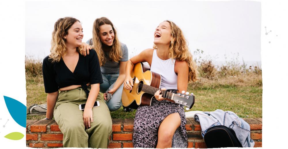 Trzy młode kobiety, jedna trzyma gitarę i śpiewa.